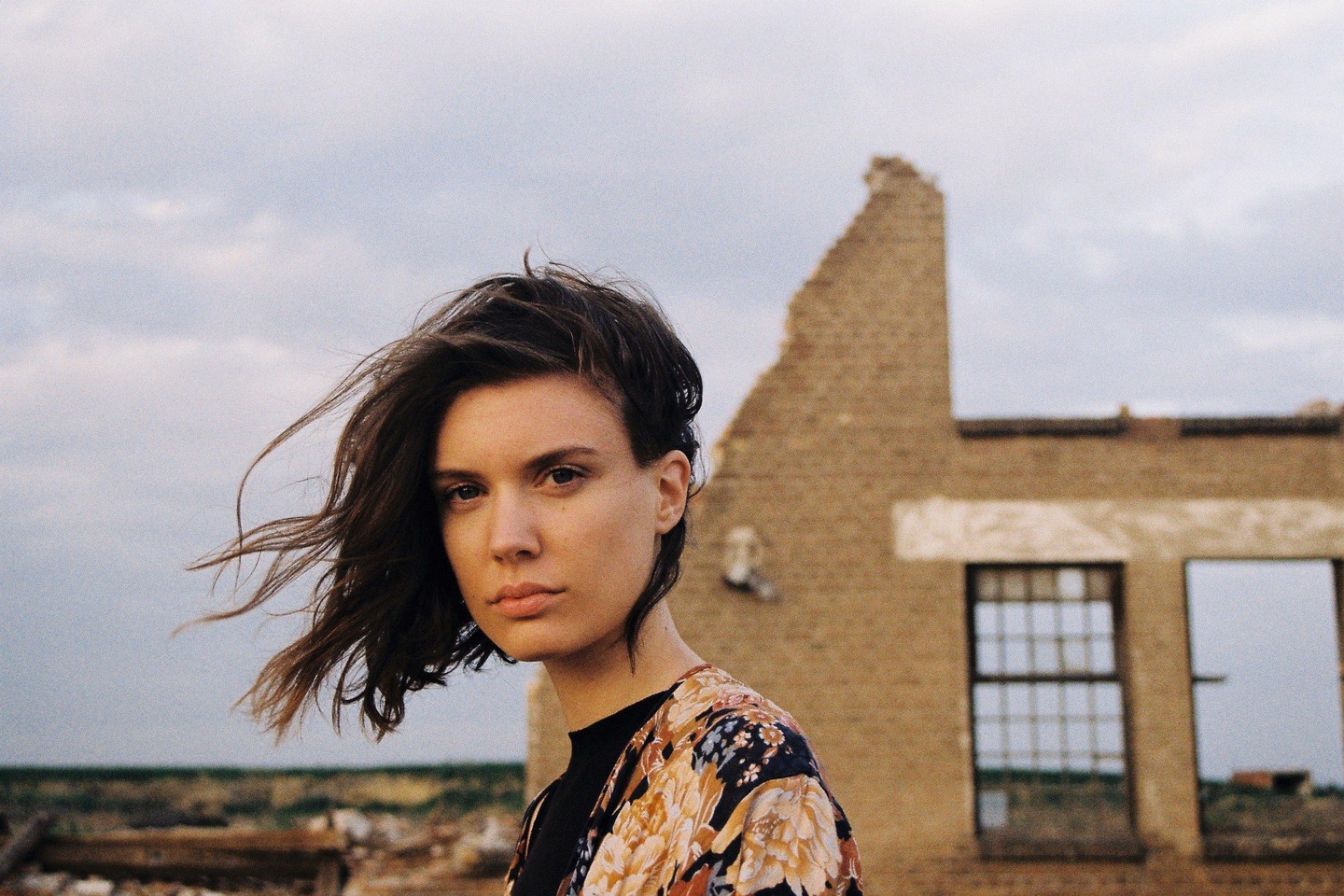 A woman with windblown hair stands in front of a ruined building.