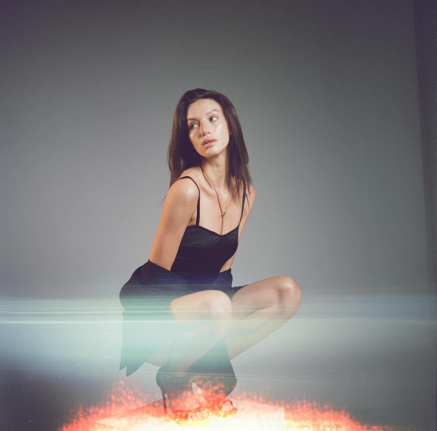A woman in a black dress and high heels crouches on a reflective surface with a light leak and red light.