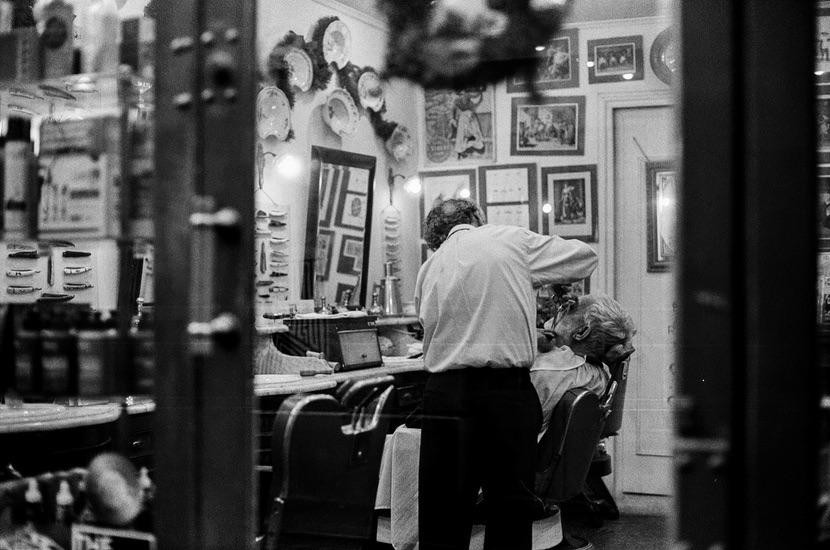 A barber shaves a man in a barbershop with many framed pictures on the wall.