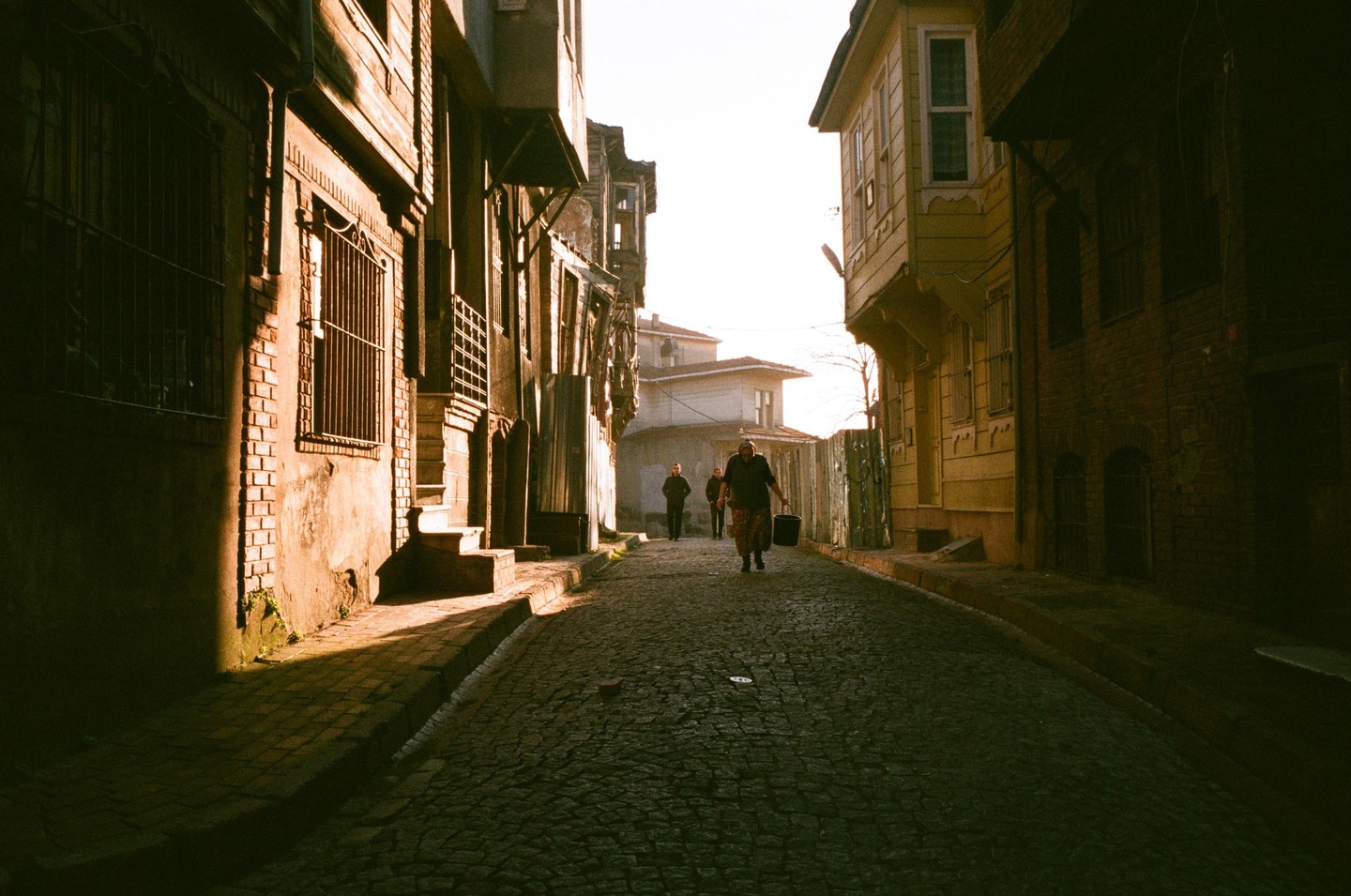 A woman carries a bucket down a cobblestone street lined with old buildings, with two men walking in the distance.