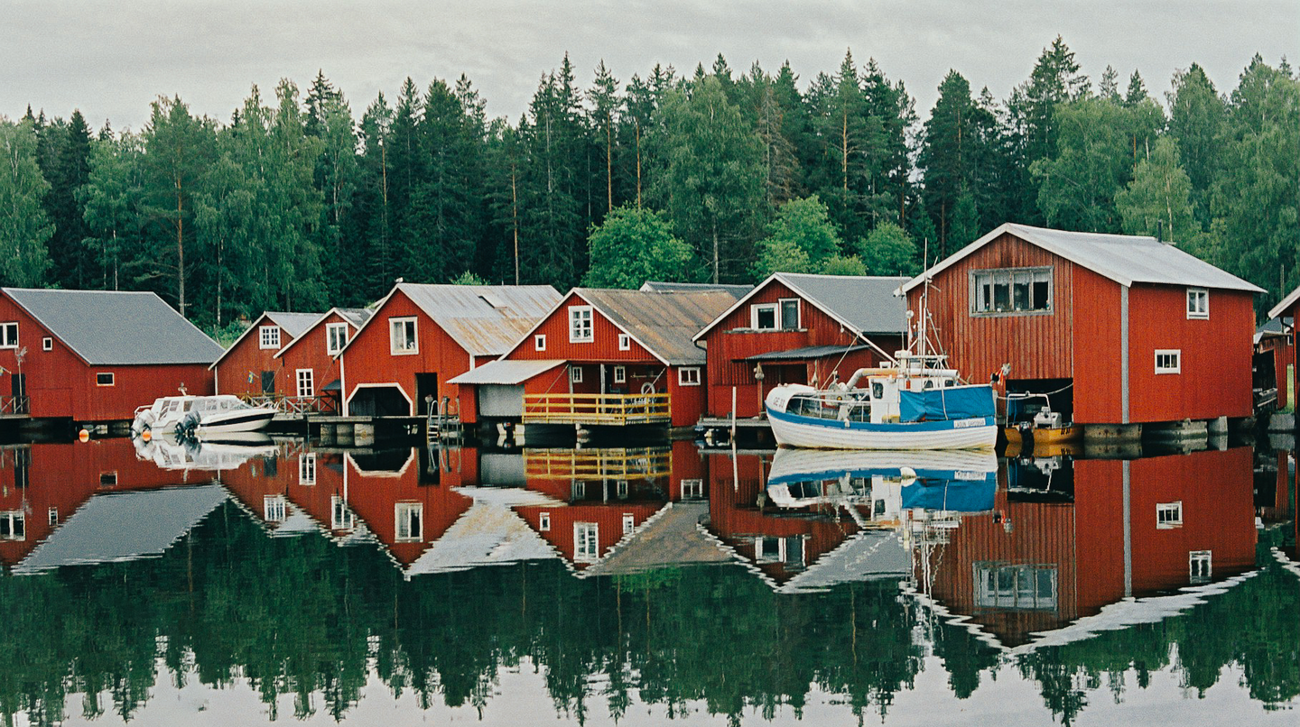 Red boathouses with white trim line a calm body of water, reflecting in the surface, with a dense forest in the background.