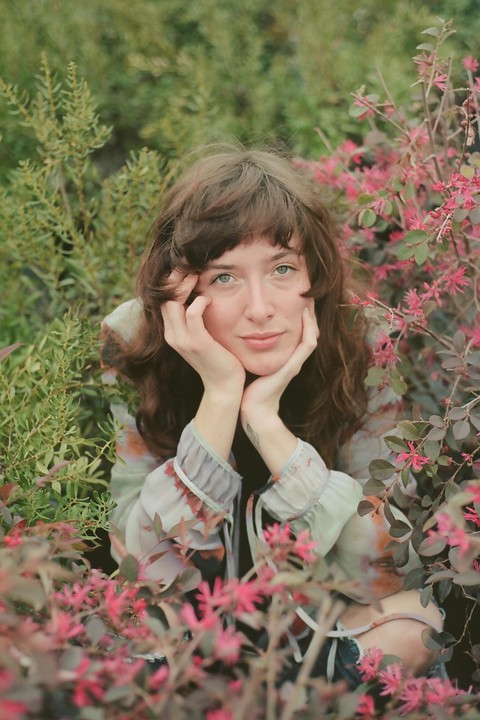 A woman with her chin resting on her hands is surrounded by green and pink foliage.