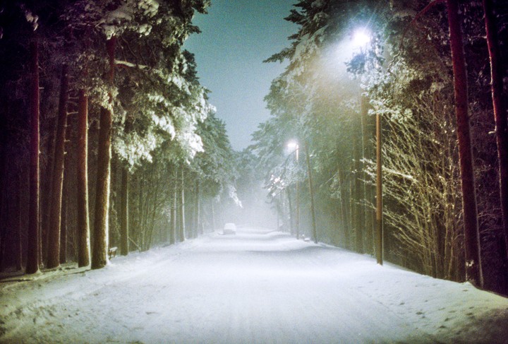 A snow-covered road is lined with trees and illuminated by streetlights at night.
