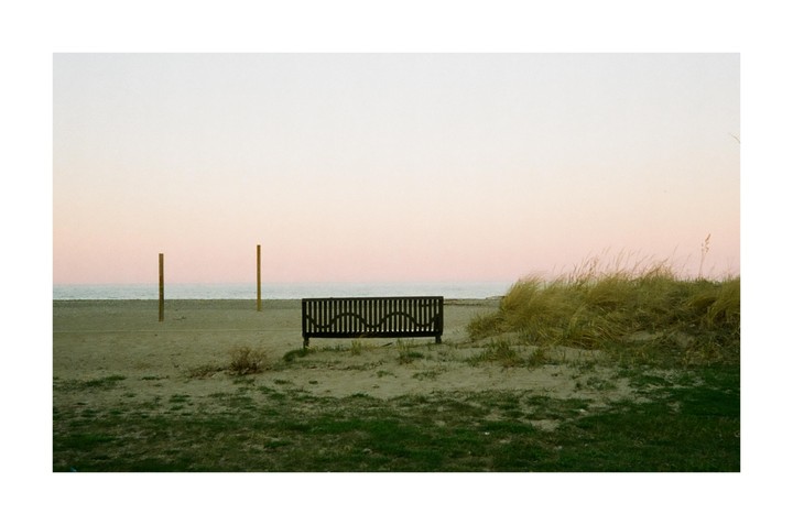 A wooden bench sits on a sandy beach with two posts and tall grass under a pink sky.