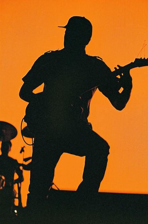 A musician in a cap plays a guitar on stage, silhouetted against an orange background.