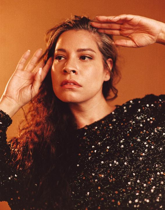 A woman with long curly hair and a sequin top poses with her hands near her head.