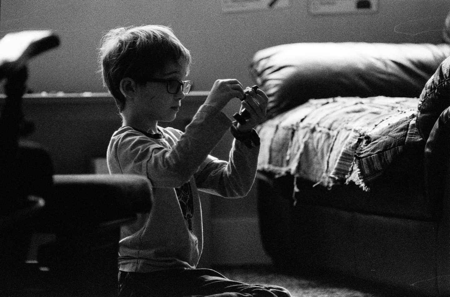 A young boy wearing glasses sits on the floor, holding and looking at a small toy.