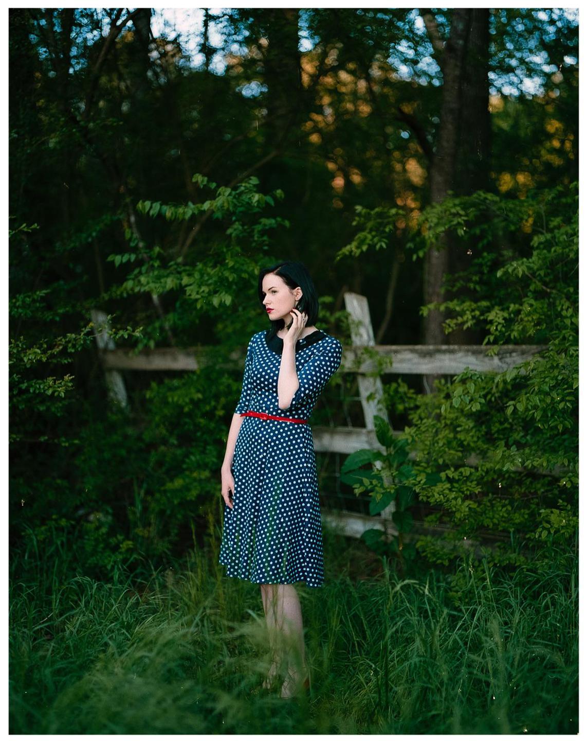 A woman in a polka dot dress stands in tall grass next to a wooden fence.