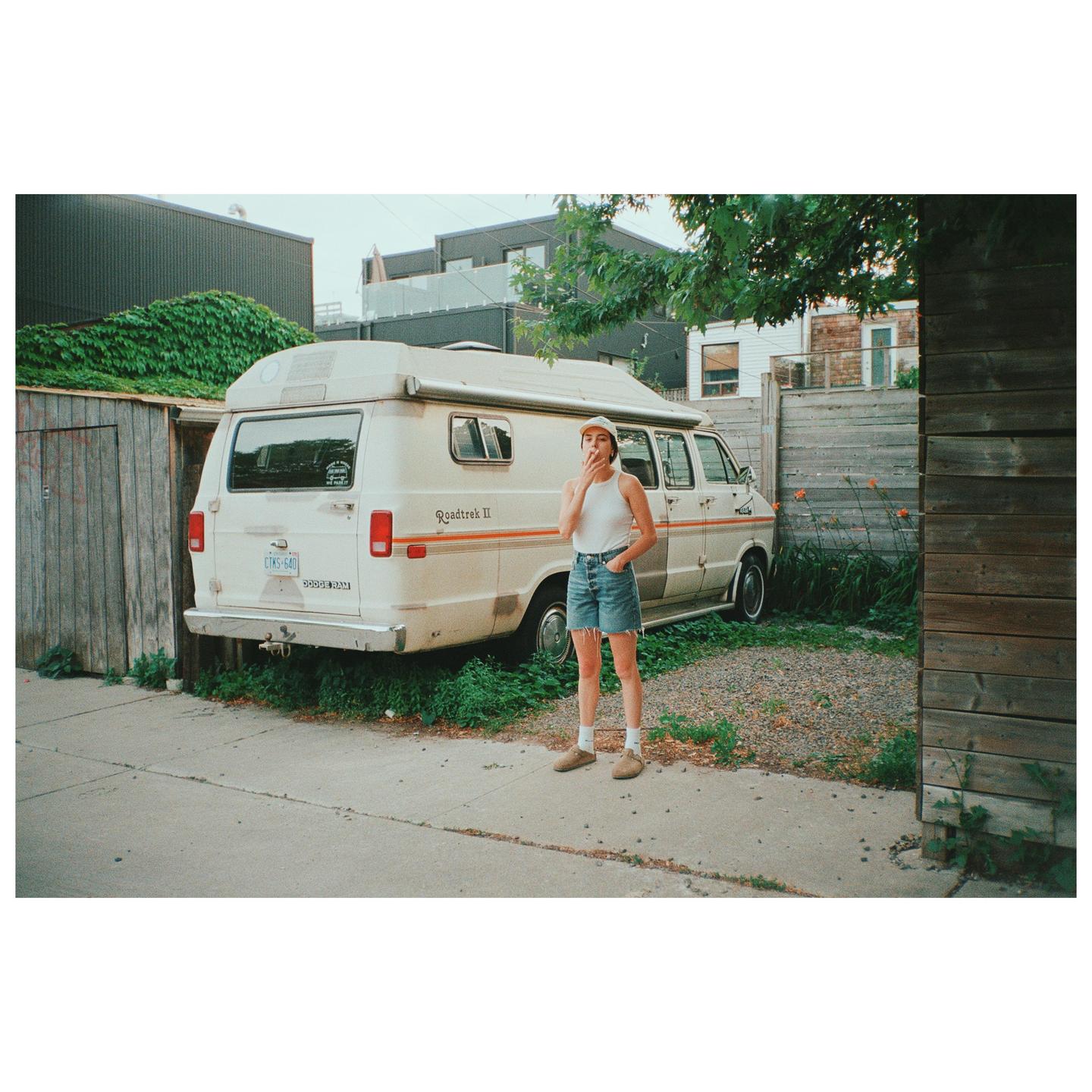 A woman stands on a sidewalk next to a parked camper van, smoking.