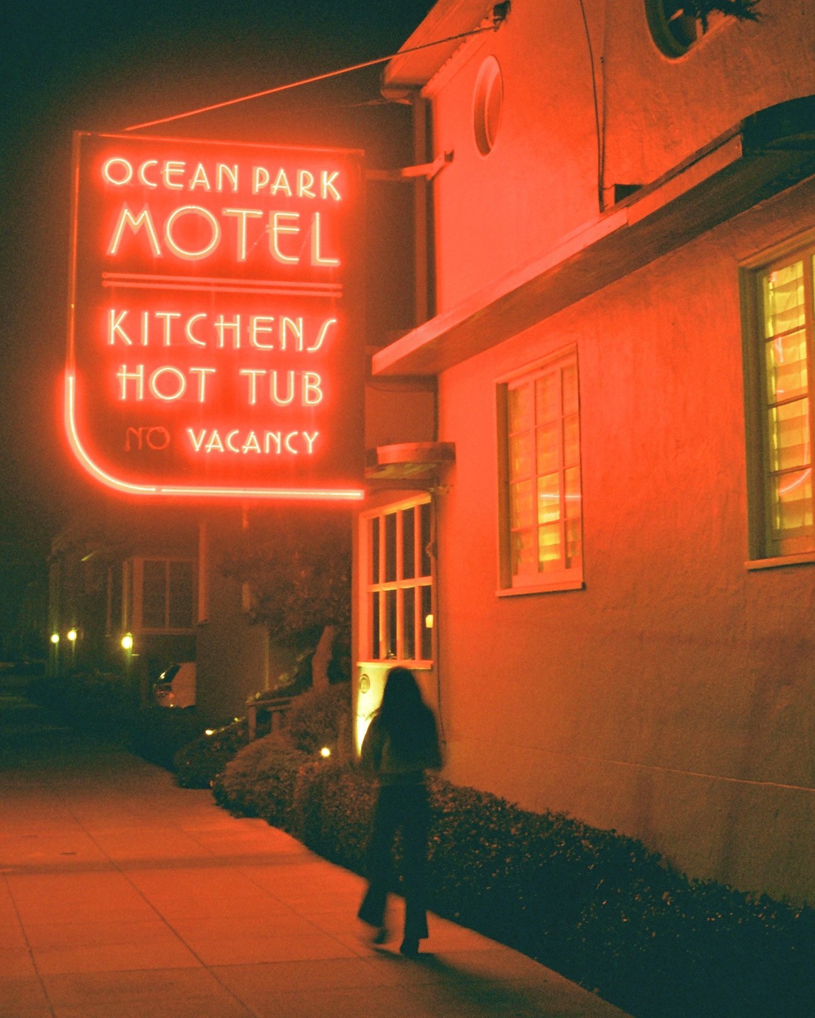 A person walks past a motel with a neon sign that reads "Ocean Park Motel, Kitchens, Hot Tub, No Vacancy" at night.