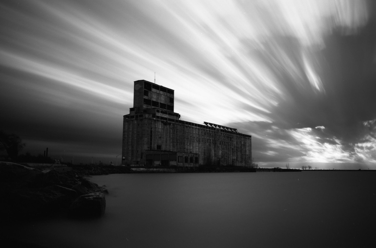 An abandoned grain elevator stands beside a body of water under a sky with streaky clouds.