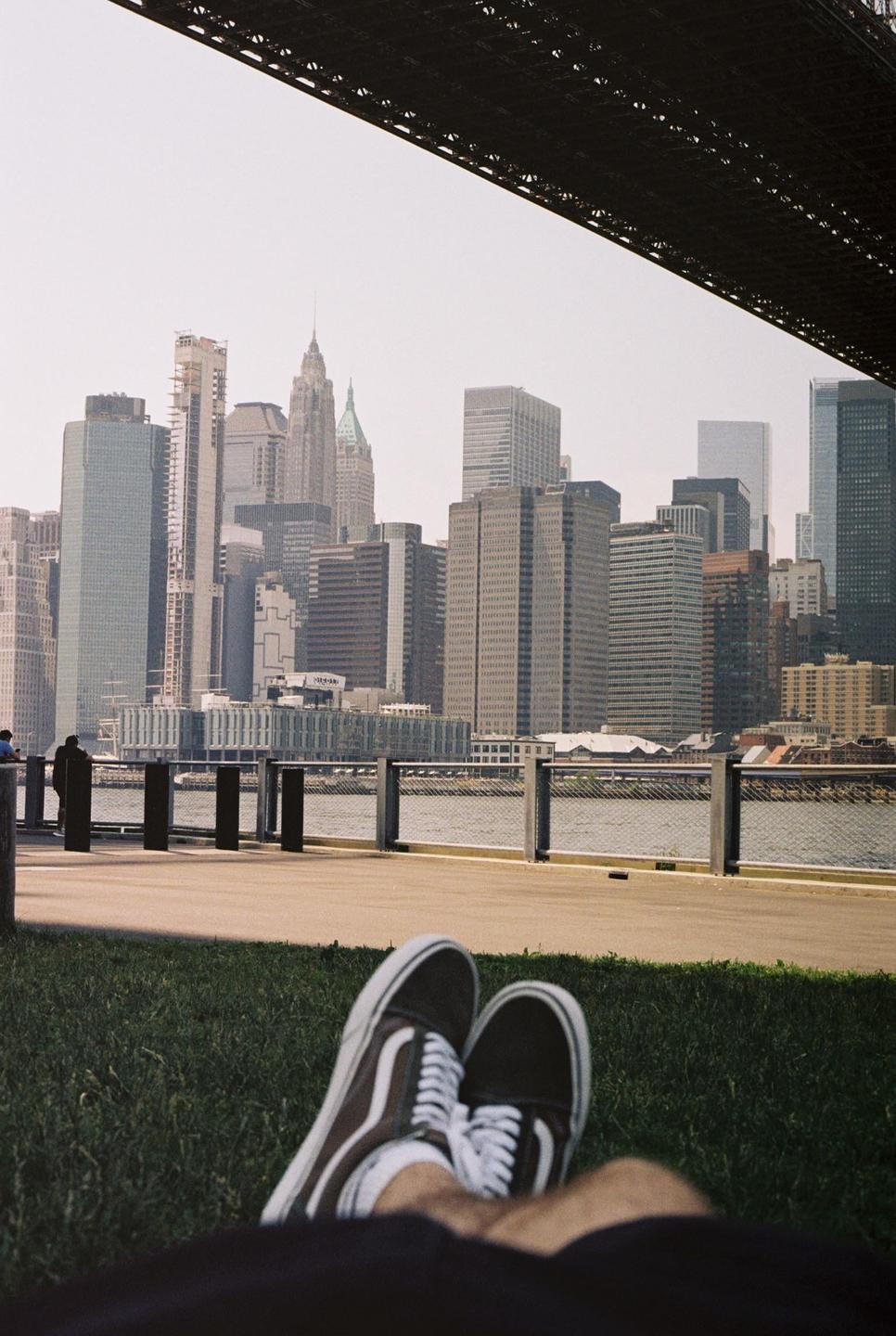 A person's feet wearing sneakers are visible in the foreground, with a city skyline and water in the background, framed by the underside of a bridge.