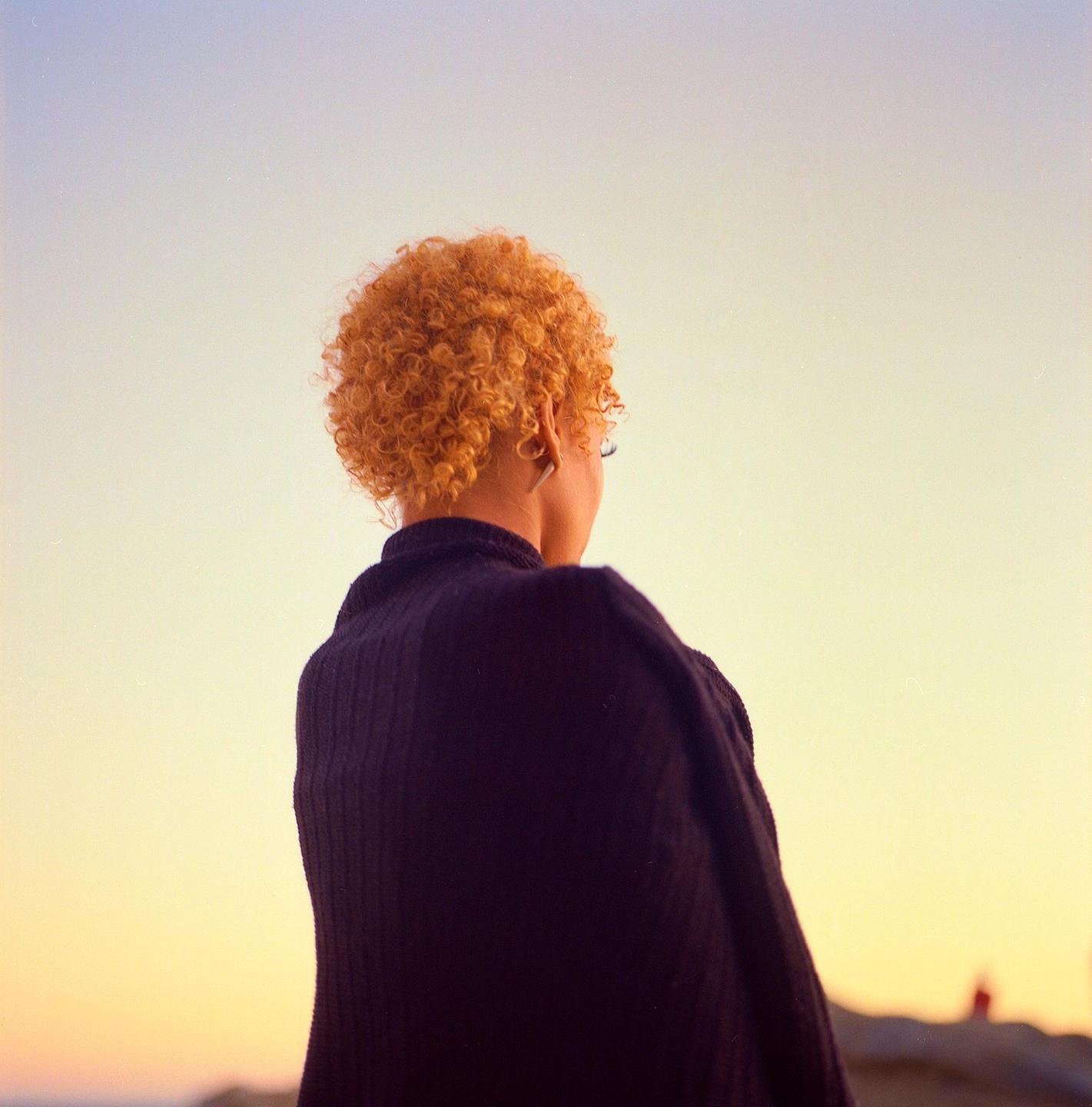A woman with curly orange hair looks out at the sunset.