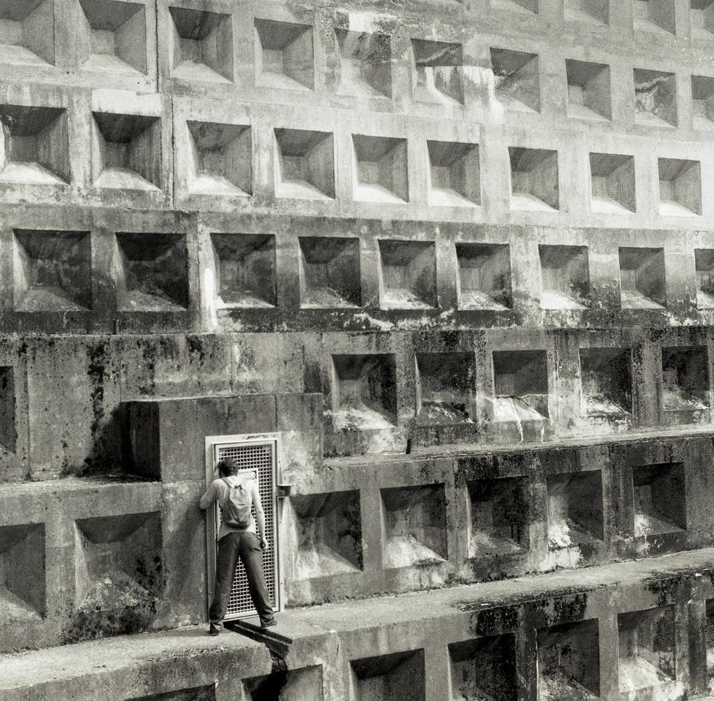 A man with a backpack stands in front of a metal door set into a concrete wall with many square indentations.