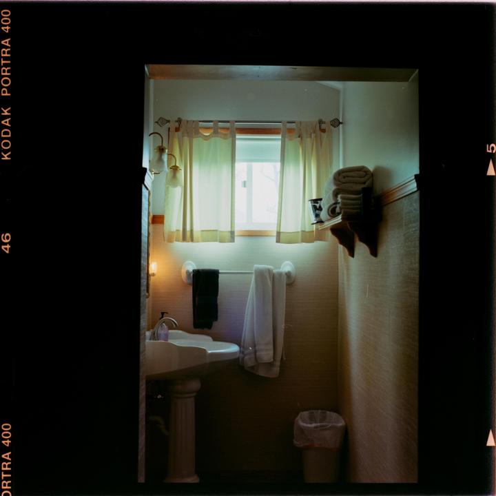 A bathroom with a pedestal sink, a window with curtains, and towels on a rack and shelf is illuminated by natural light and sconces.
