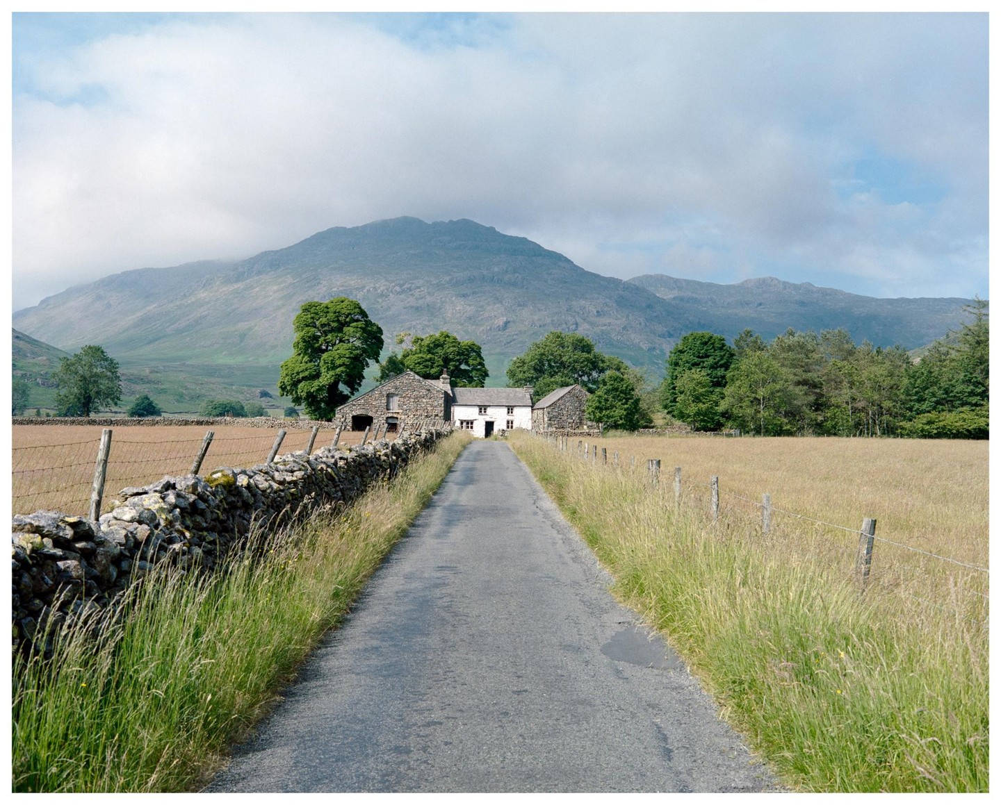 A narrow road leads to a white house and stone barn in a valley with mountains in the background.