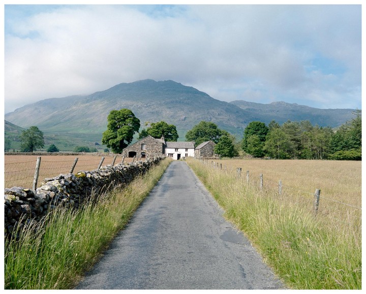 A narrow road leads to a white house and stone barn in a valley with mountains in the background.