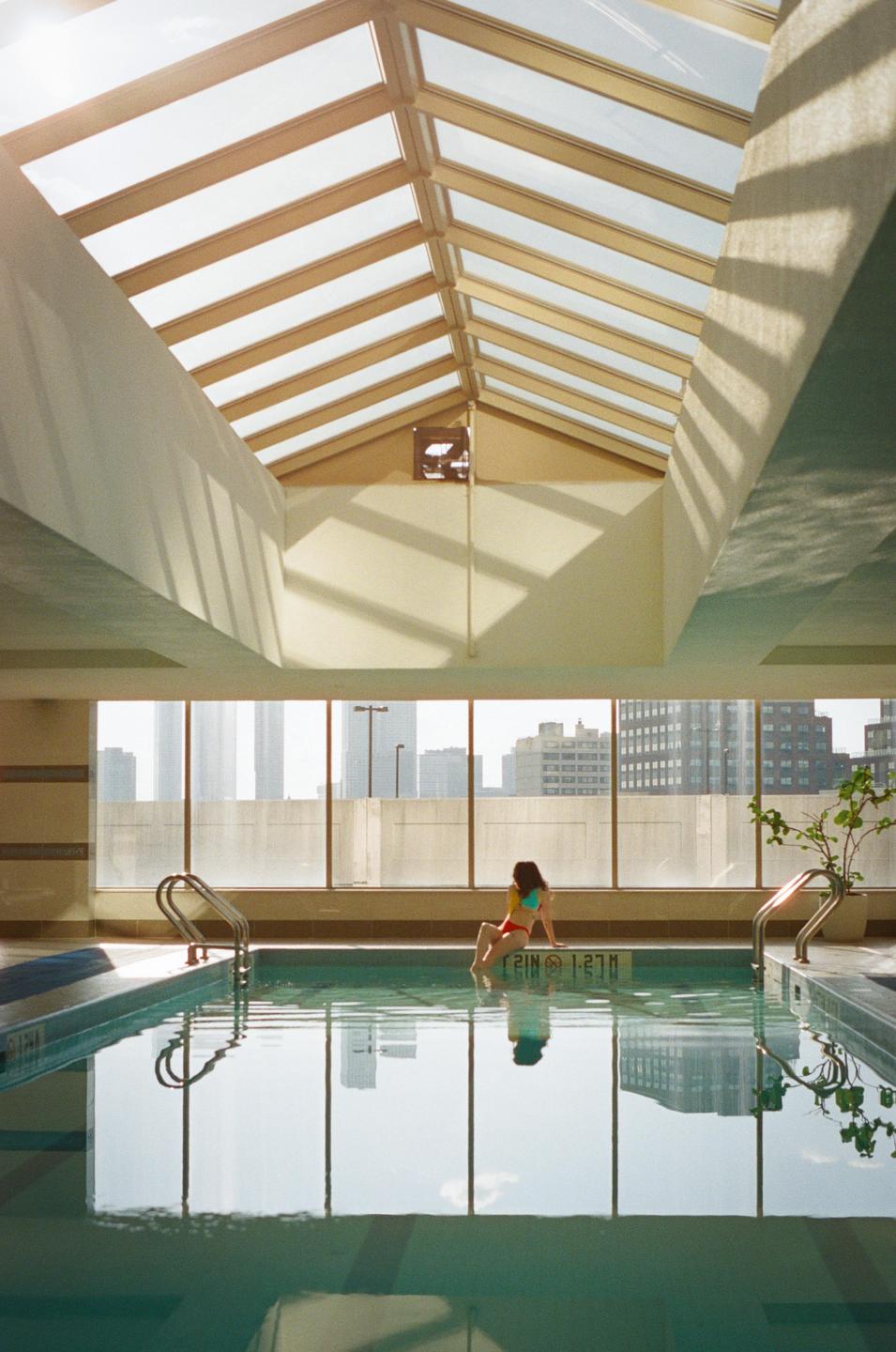 A woman sits on the edge of an indoor swimming pool with a city skyline visible through large windows.