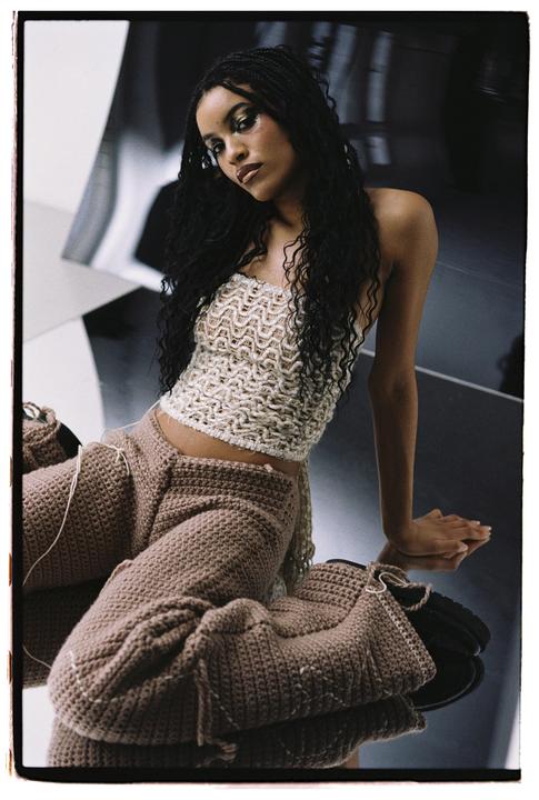 A woman with long, dark, braided hair and glittery eye makeup sits wearing a crocheted top and pants.