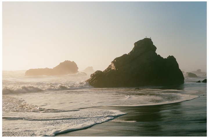 Waves crash against large rocks on a hazy beach.