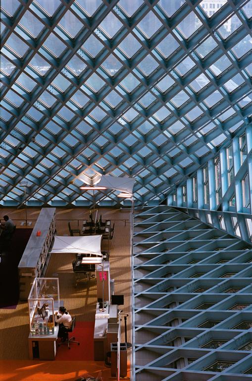 A woman works at a desk under a large glass and metal grid ceiling in a modern library.