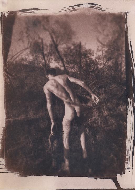 A man stands naked in a marsh, his back to the viewer, with trees and foliage in the background.