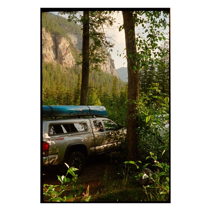 A silver pickup truck with a blue canoe on its roof is parked among trees with a mountain in the background.