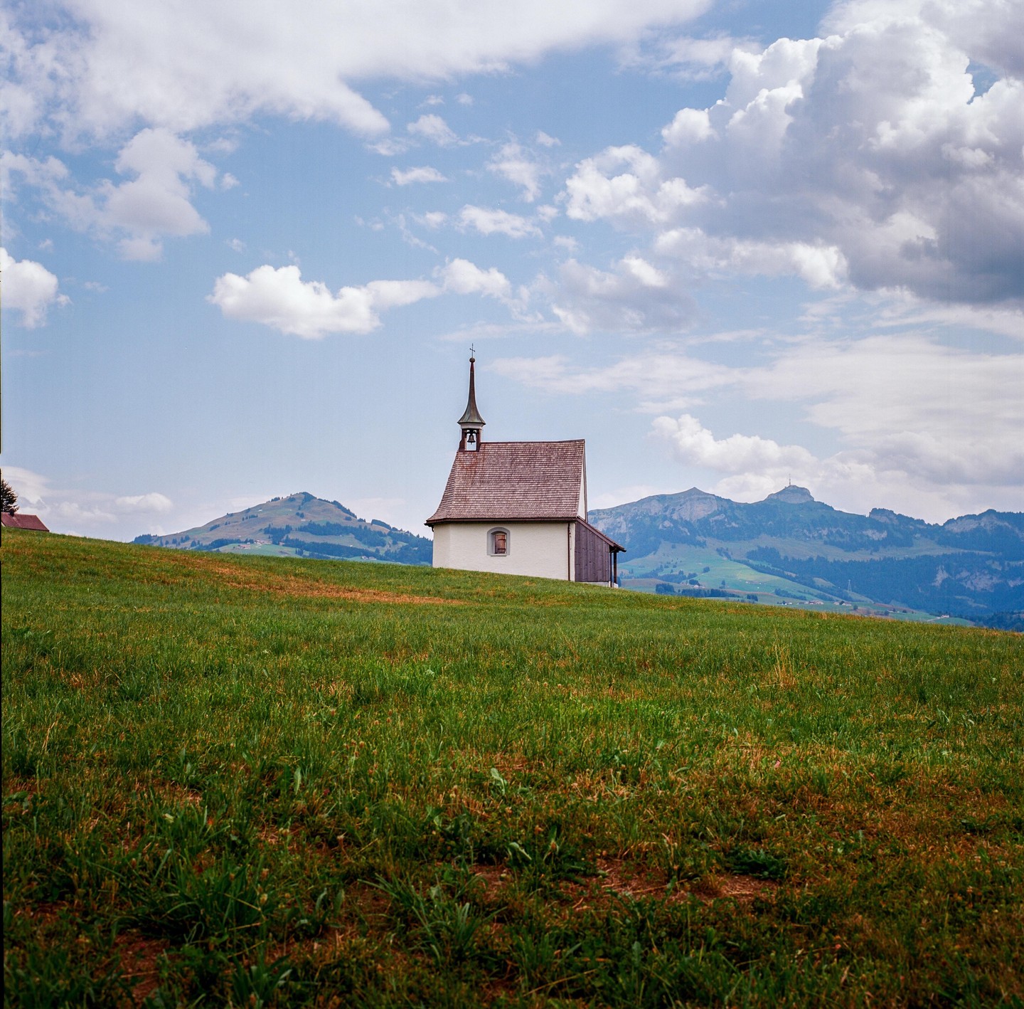 A small chapel with a steeple stands on a grassy hill with mountains in the background under a cloudy sky.