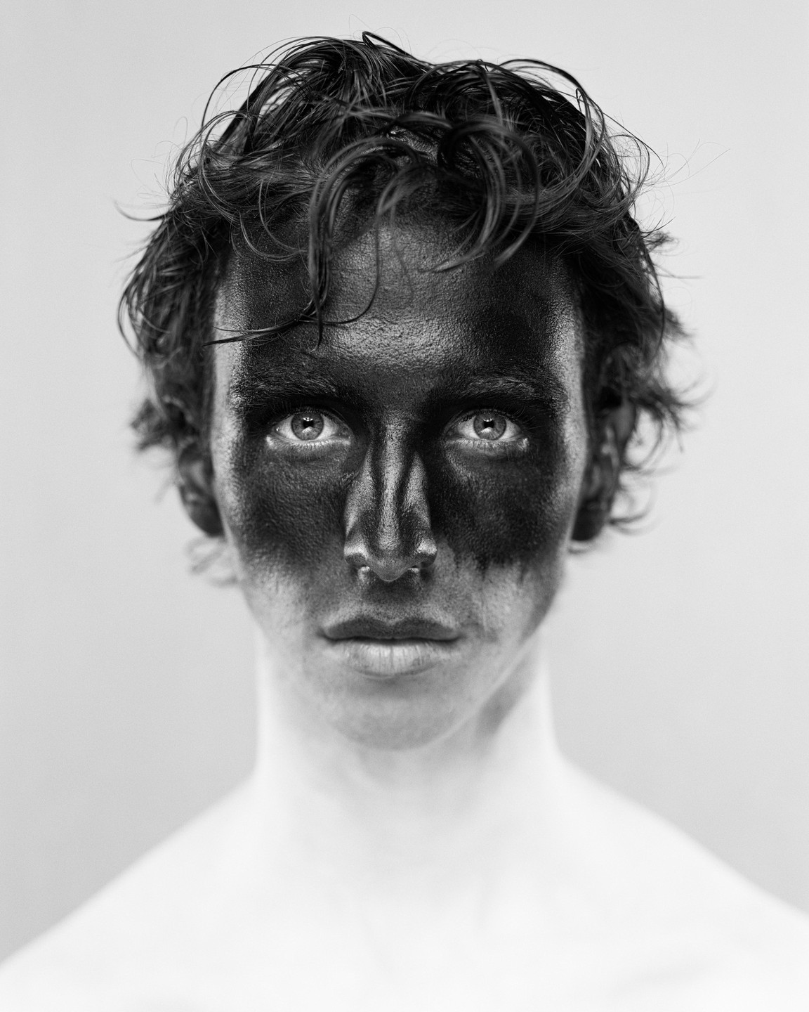 A man with black paint covering his face looks directly at the viewer.