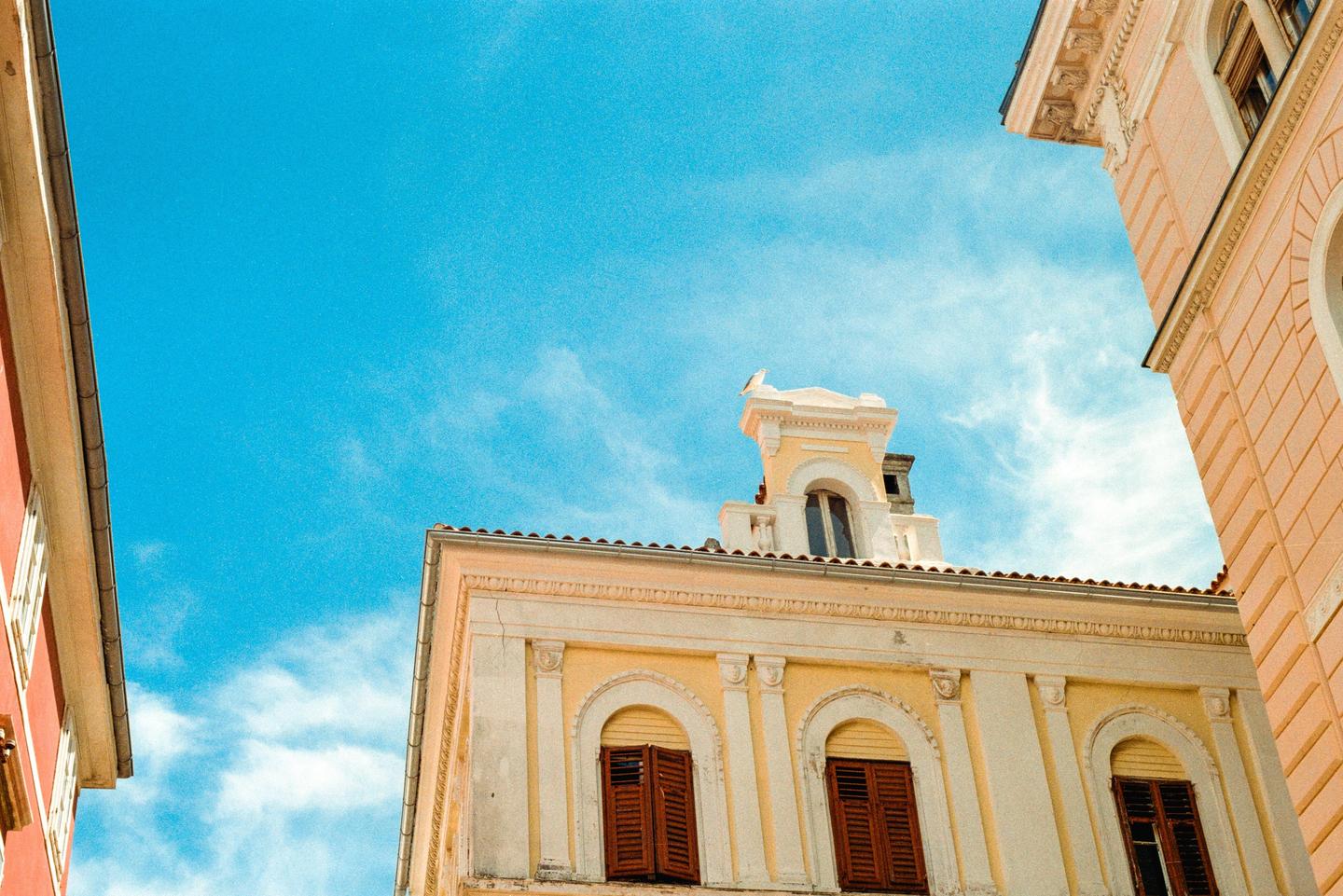 A low-angle view shows the facades and rooftops of several buildings against a bright blue sky with scattered clouds.