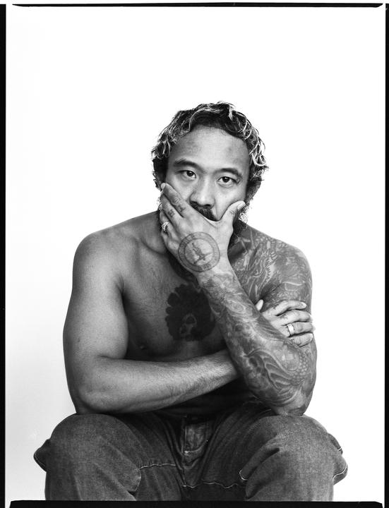 A man with tattoos on his chest and arms sits with his hand covering his mouth.