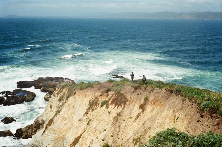 Two men stand on a cliff overlooking a vast ocean with waves crashing against the shore.