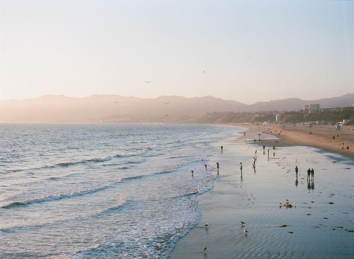 A wide view of a beach with people walking along the shore and mountains in the distance under a hazy sky.