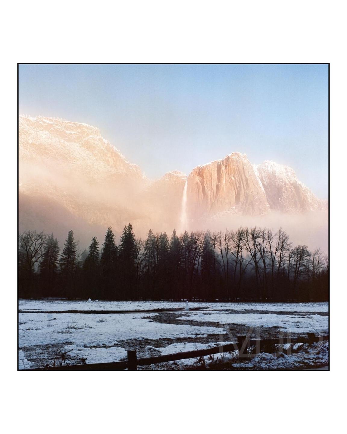 A waterfall cascades down a mountain face, partially obscured by mist, with a snow-covered field and forest in the foreground.