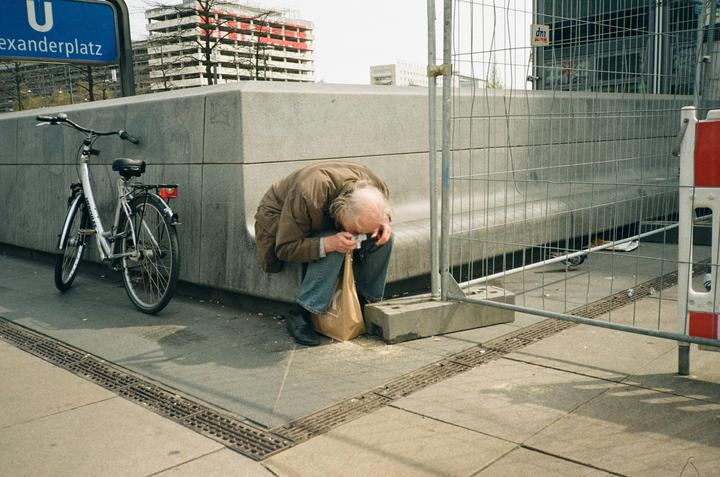 A man bends over a paper bag on the ground next to a bicycle and a construction fence.