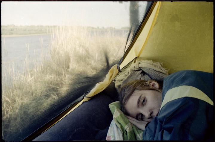 A woman lies inside a tent looking out at a body of water and tall grass.