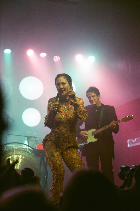 A woman in a floral jumpsuit sings into a microphone on stage while a man plays a guitar in the background.