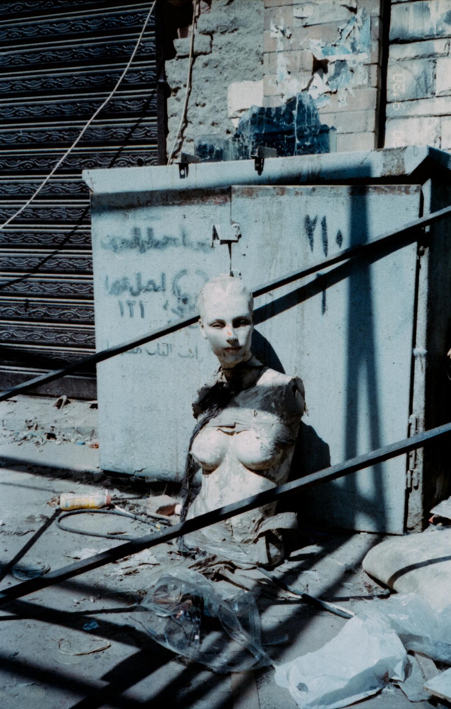 A broken mannequin torso with a head is on the ground in front of a metal box with Arabic writing.