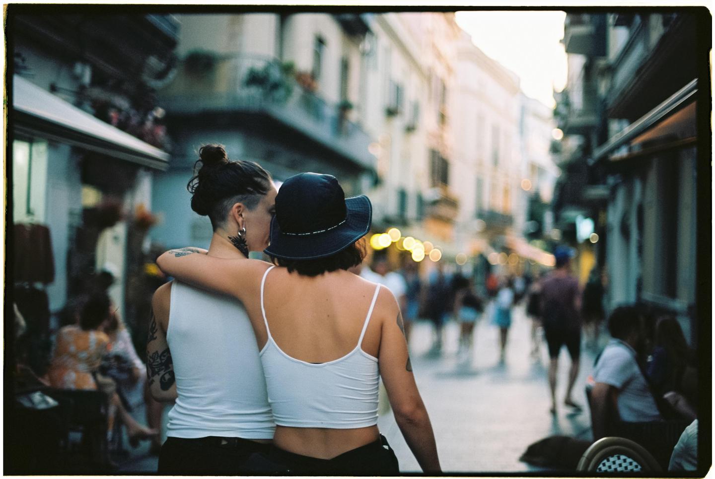 Two women embrace on a busy street at dusk, with buildings and lights blurring in the background.