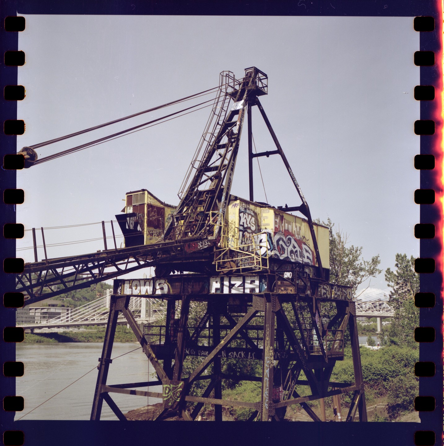 An old, rusty, yellow crane covered in graffiti stands by a river with a bridge in the background.