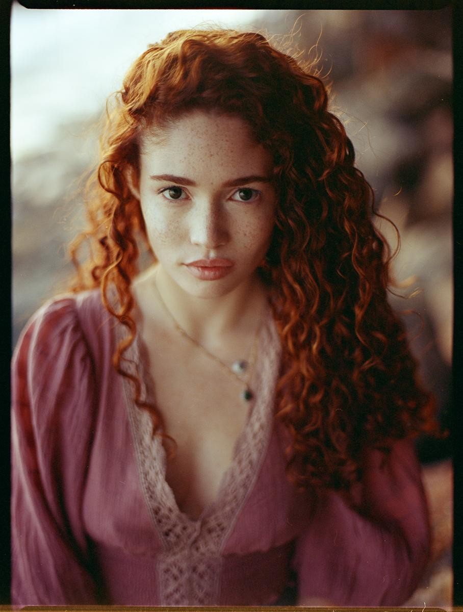A woman with long, curly red hair and freckles looks directly at the viewer.