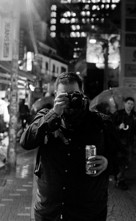 A man holds a camera to his eye and a can in his other hand on a city street at night.