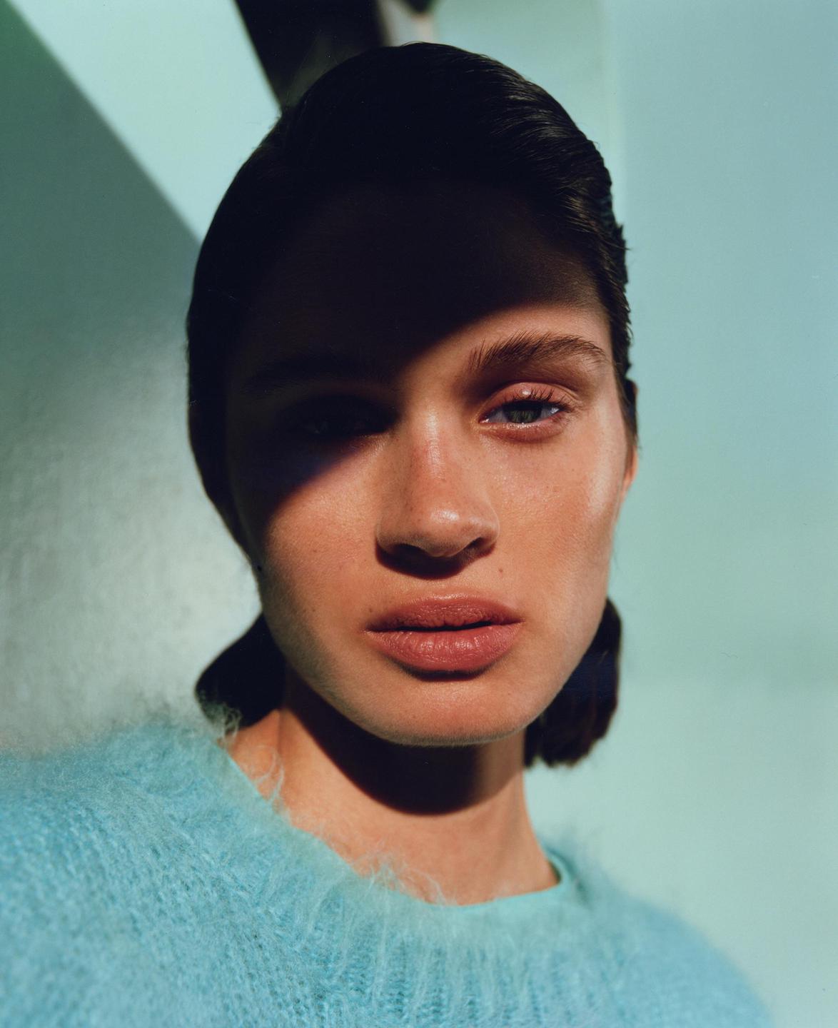 A woman wearing a blue sweater is illuminated by a beam of light that casts a shadow across her face.