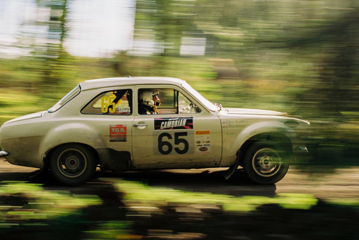 A white rally car with the number 65 on its side speeds past, blurring the green trees in the background.