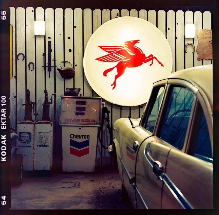 A vintage car is parked next to old gas pumps and a large illuminated sign with a red Pegasus logo.
