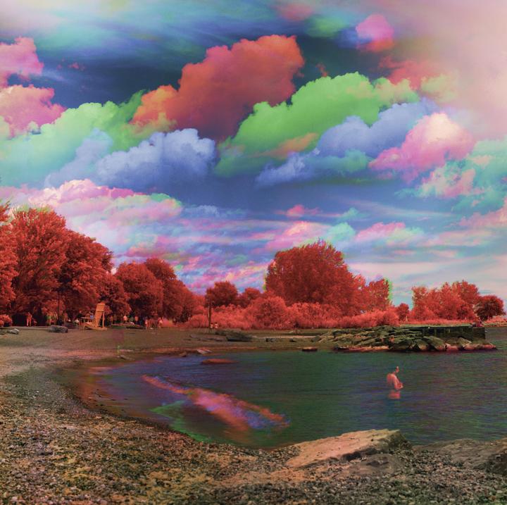 A man stands in the water at a rocky beach with colorful clouds in the sky.