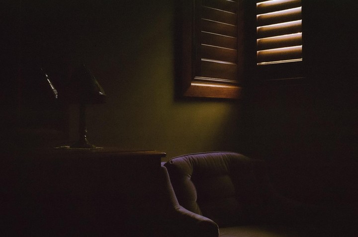 Sunlight streams through a window with wooden blinds, illuminating a lamp and a chair in a dimly lit room.