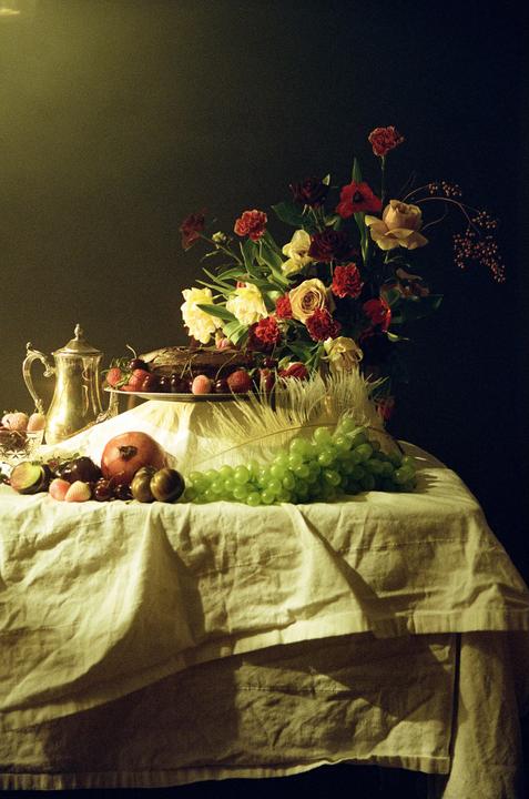 A still life arrangement features a bouquet of flowers, a cake, fruit, and a silver pitcher on a draped table.