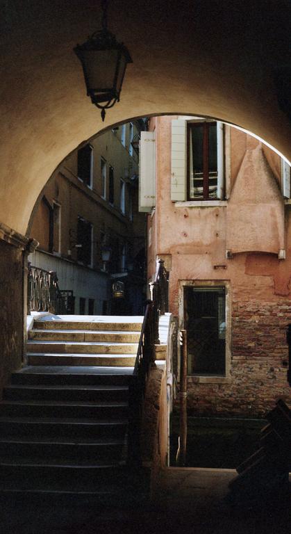 A dark archway with a hanging lantern opens to a sunlit street with stairs and buildings in Venice.
