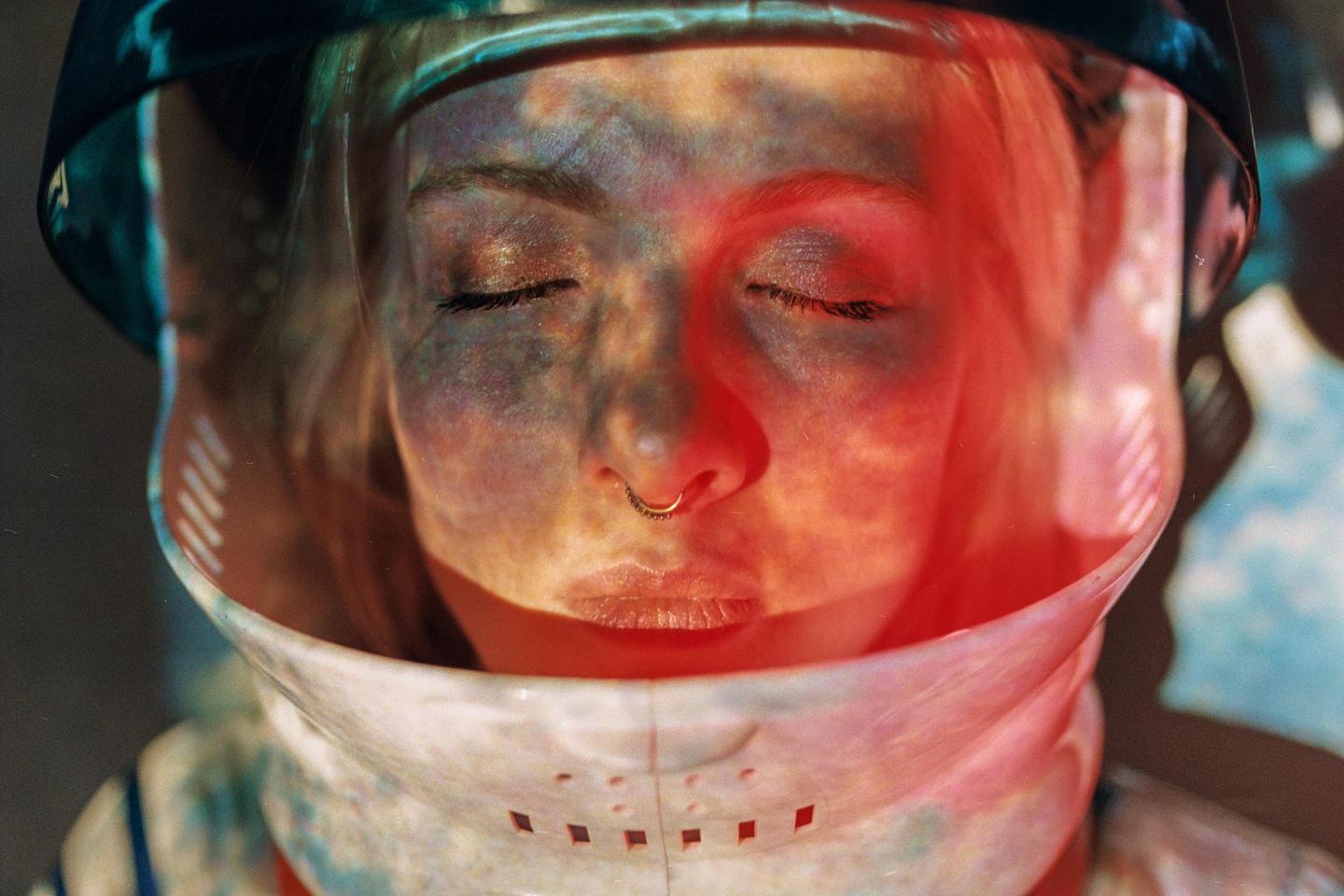 A woman with closed eyes wears a space helmet with colorful light projected onto her face.